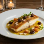 A plated pan-seared fish fillet with a crispy golden crust, drizzled with a rich sauce and garnished with dried fruit, slivered almonds, and fresh herbs.