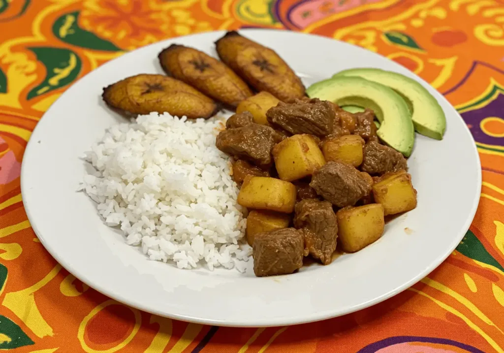 A plated meal featuring Carne Con Papas alongside white rice, fried plantains, and avocado slices, set on a vibrant, patterned tablecloth.